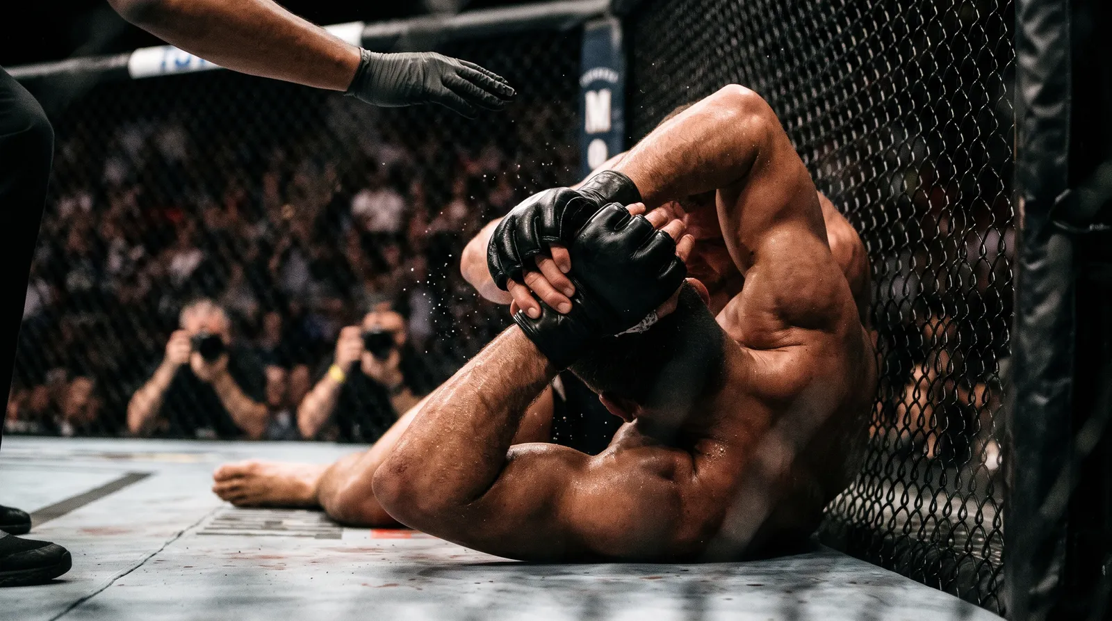 Gloved hands locked in a front choke against the octagon fence as a referee's hand reaches in