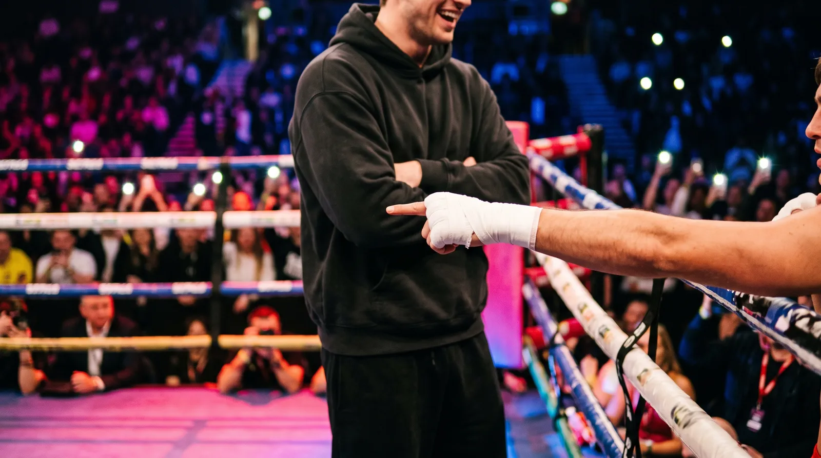 A coach's taped hand points across the ring ropes while a tall figure in a hoodie laughs behind him