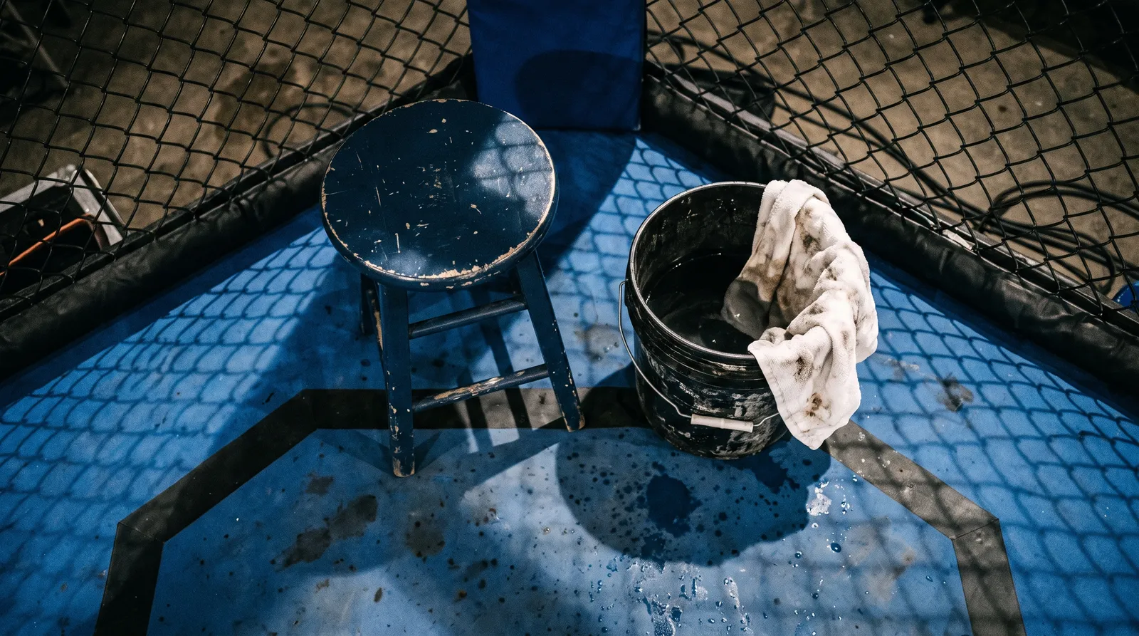 A corner stool and water bucket on the octagon canvas under bright arena lights, with the fence's shadow across them