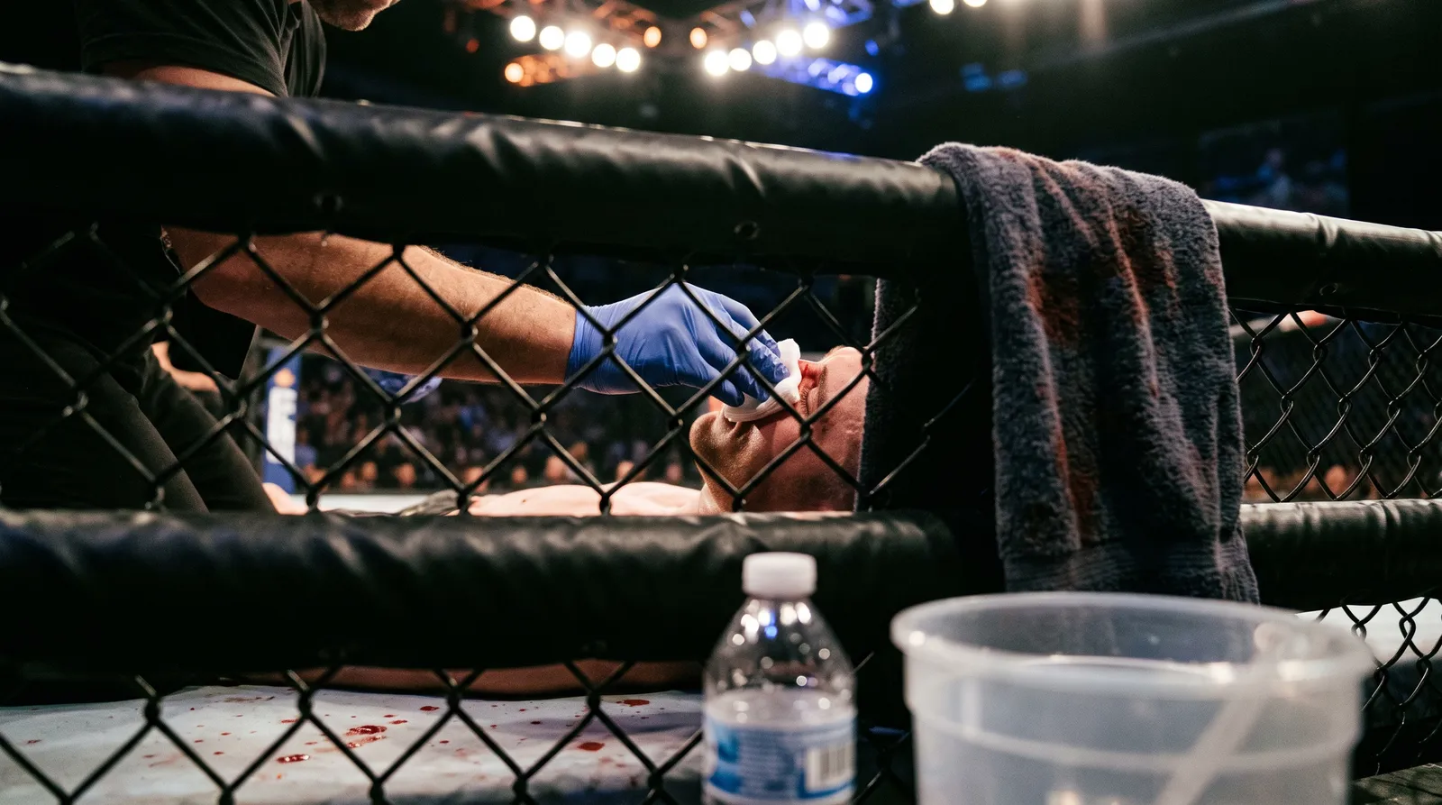 A cornerman presses gauze to a bloodied fighter's nose between rounds at the cage