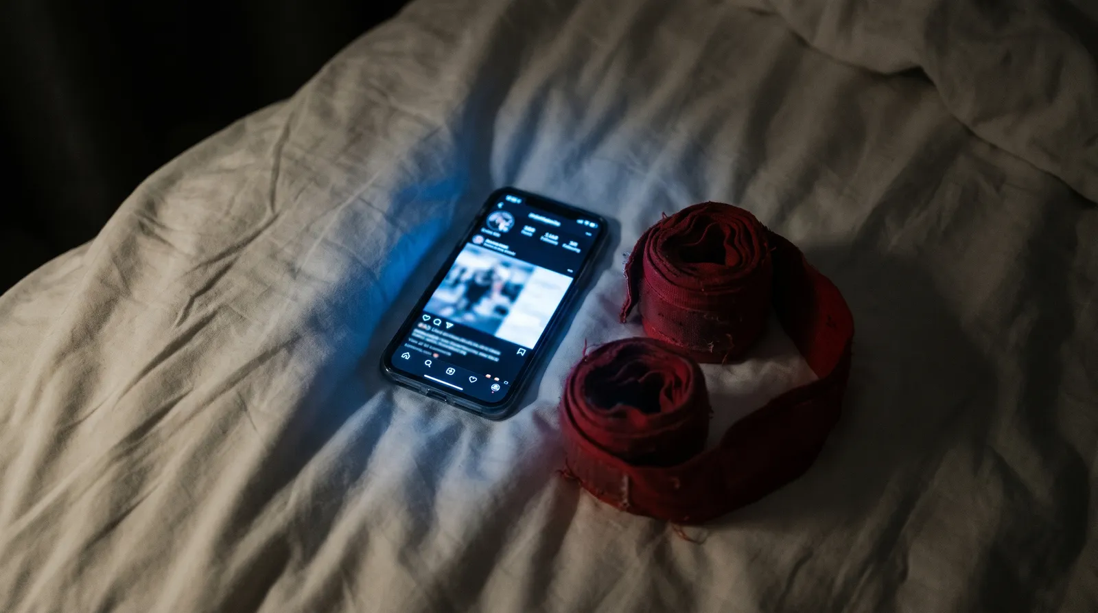 A glowing phone screen beside coiled boxing hand wraps on a dark hotel bed