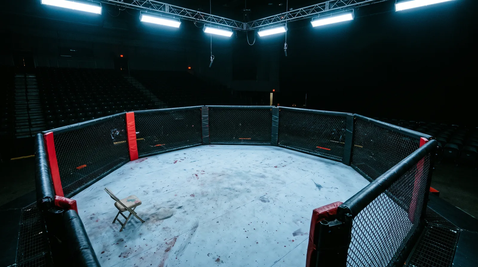 An empty UFC octagon under work lights with a lone corner stool by the fence