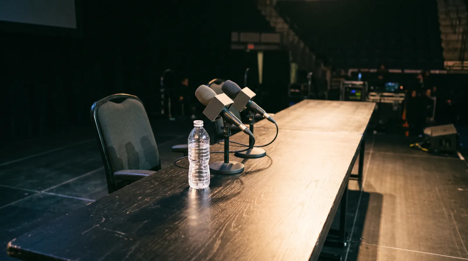An empty press conference table with microphones and a pushed-back chair under a single stage light