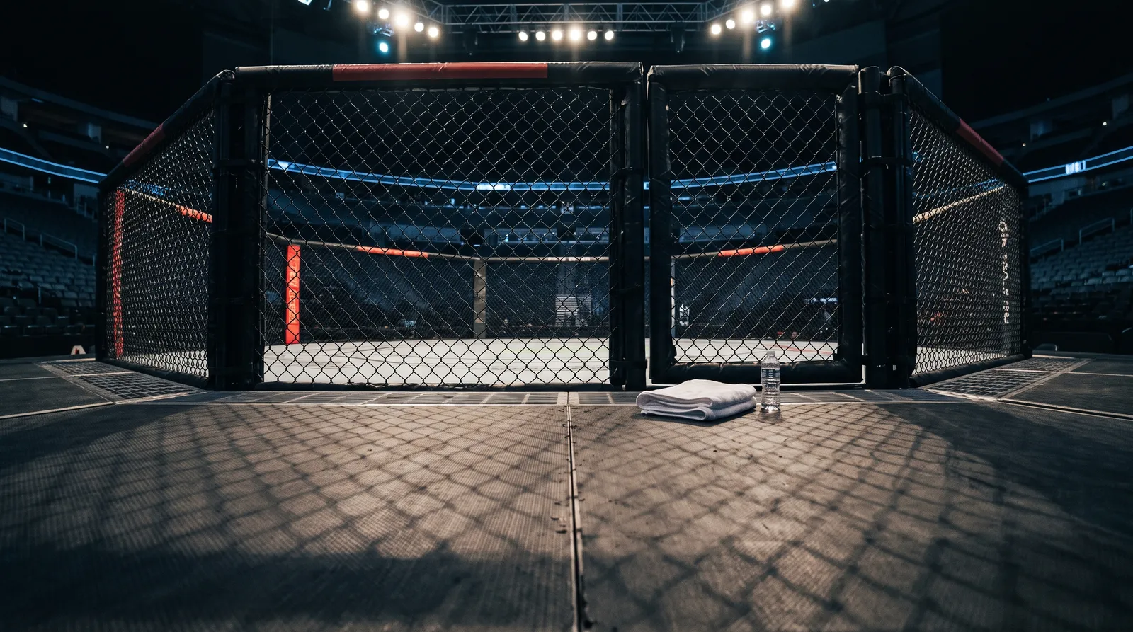An empty octagon under arena work lights with a towel and water bottle near the cage door