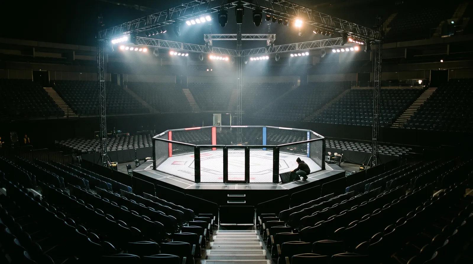 An empty octagon under work lights in a darkened arena before fight night