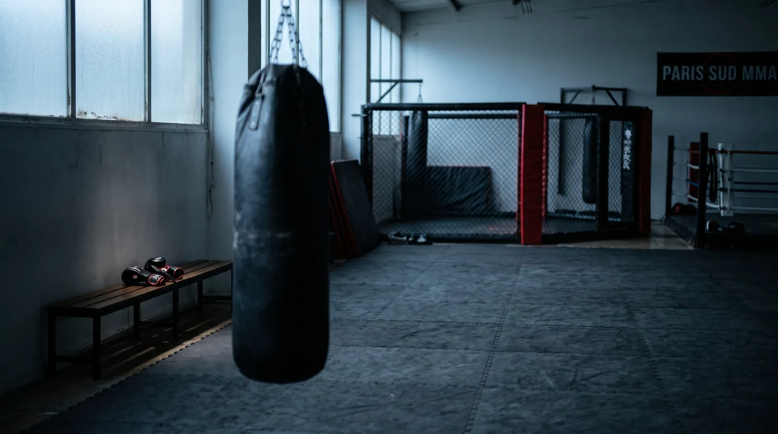 Empty MMA gym at dawn with a still heavy bag and a pair of gloves resting on a bench