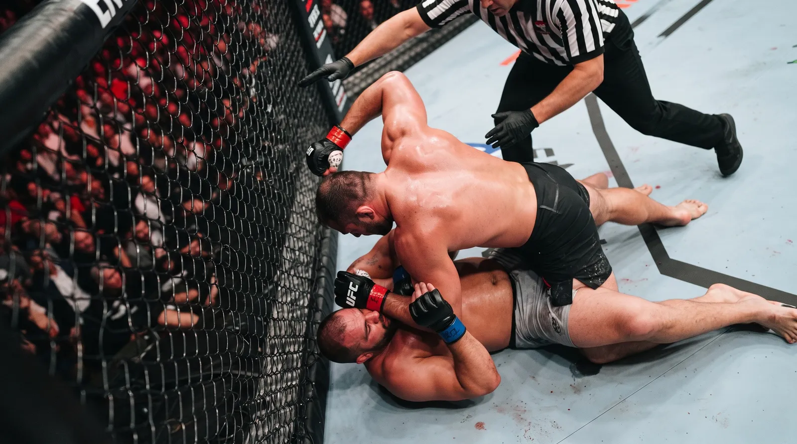 A referee reaches in as a heavyweight rains down punches on a trapped opponent against the cage