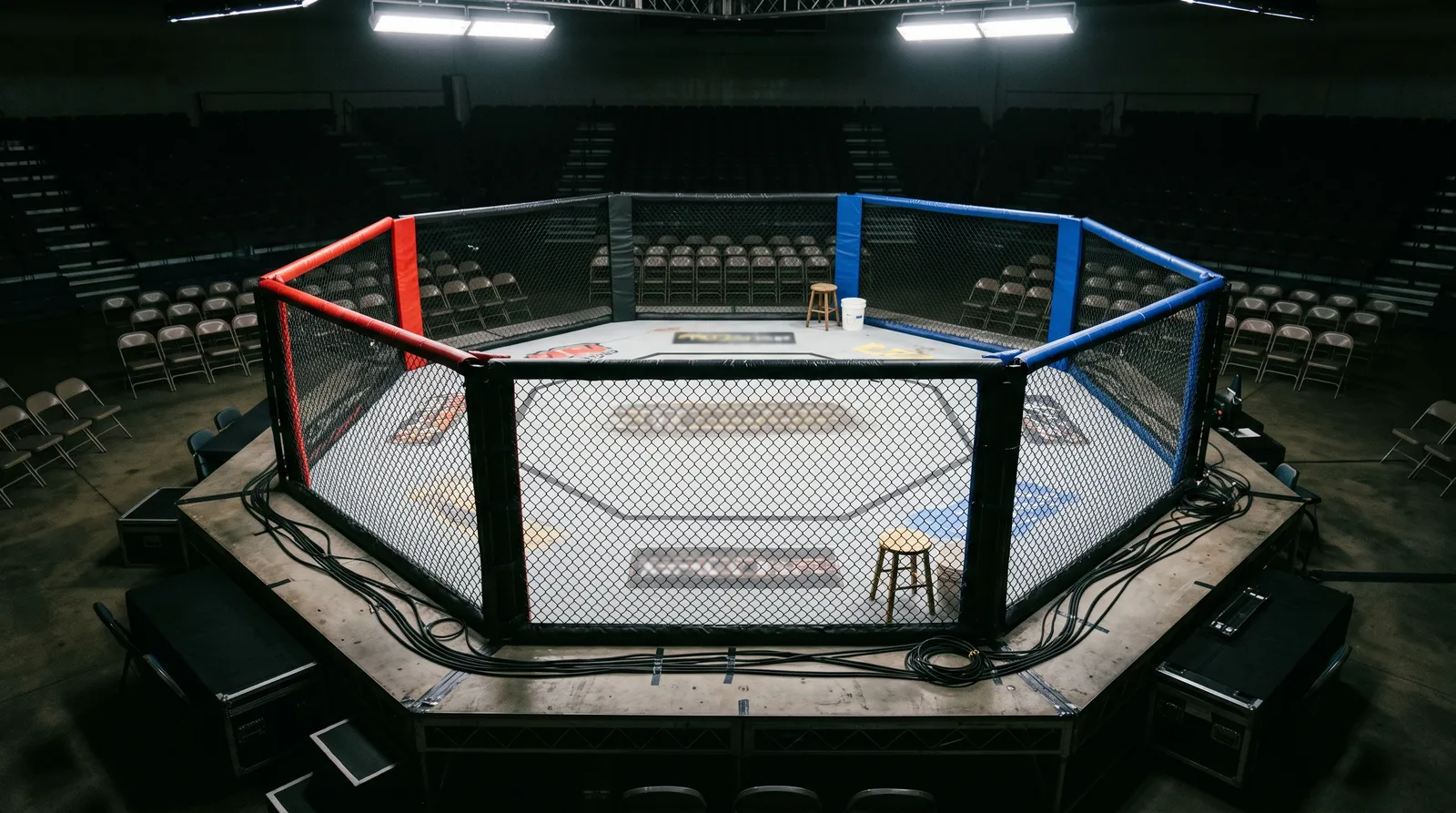 An empty octagon under bright work lights with a corner stool and bucket before fight night