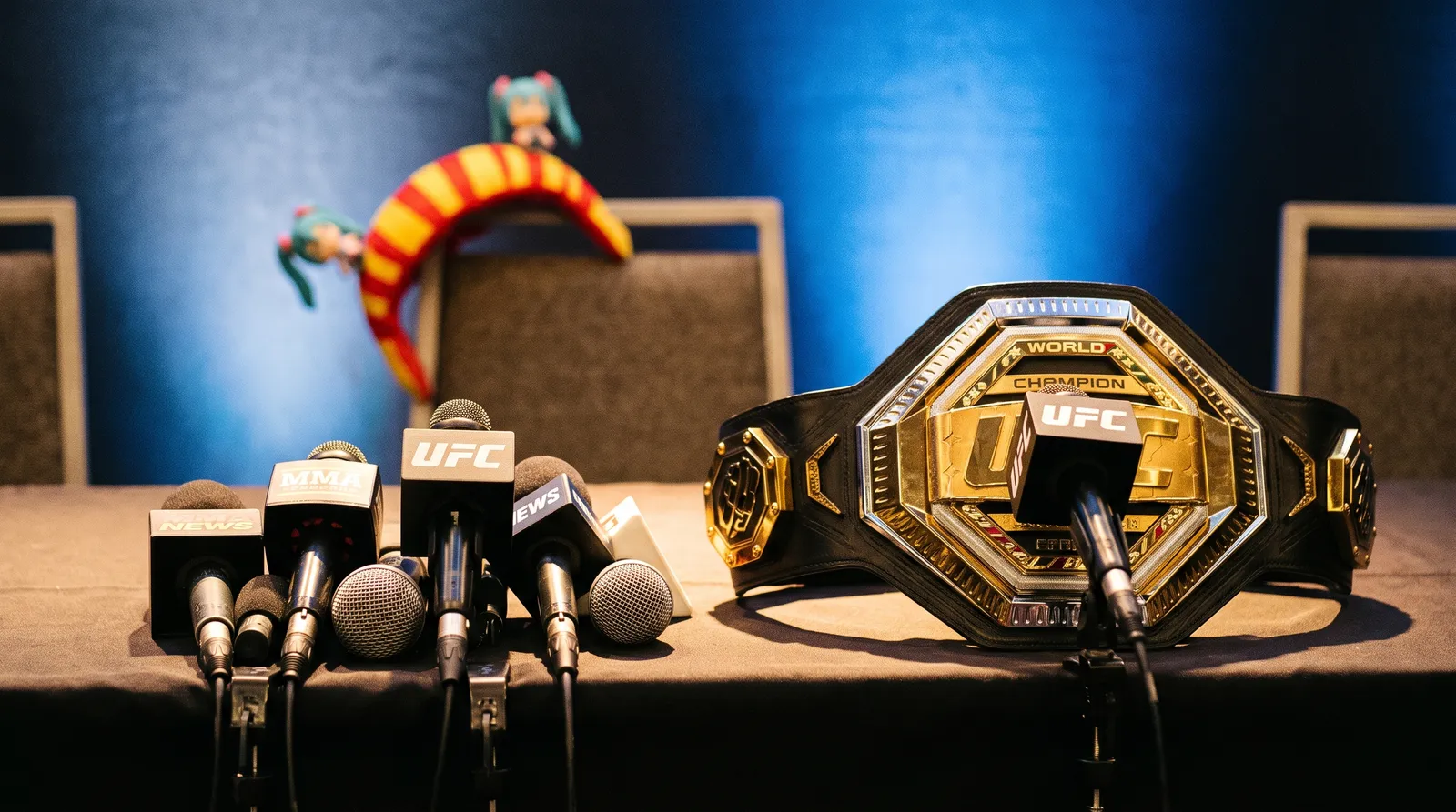 Microphones and a championship belt on a press conference table with a costume piece draped over an empty chair