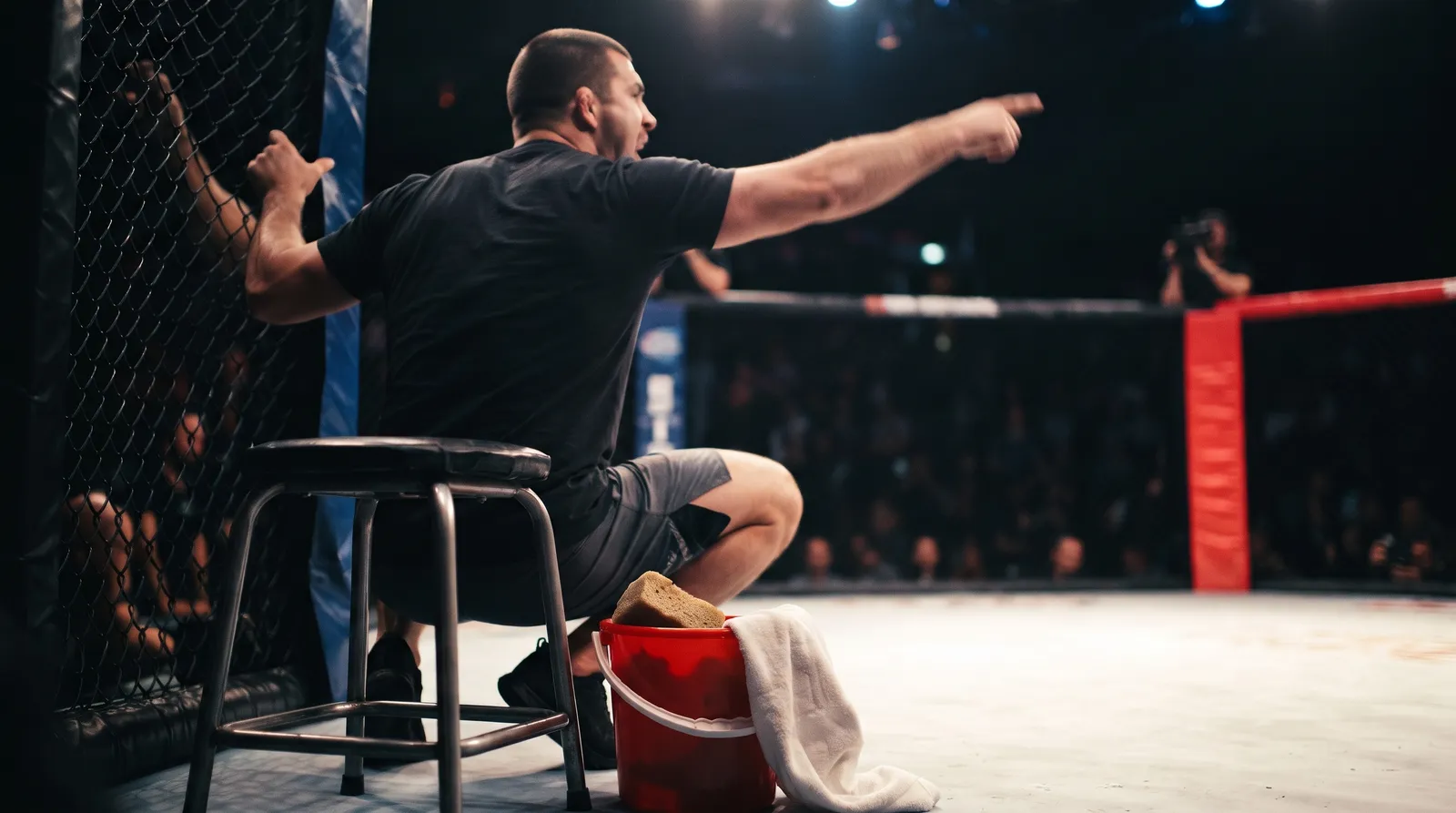 A coach yells instructions through the cage fence beside a corner stool and water bucket